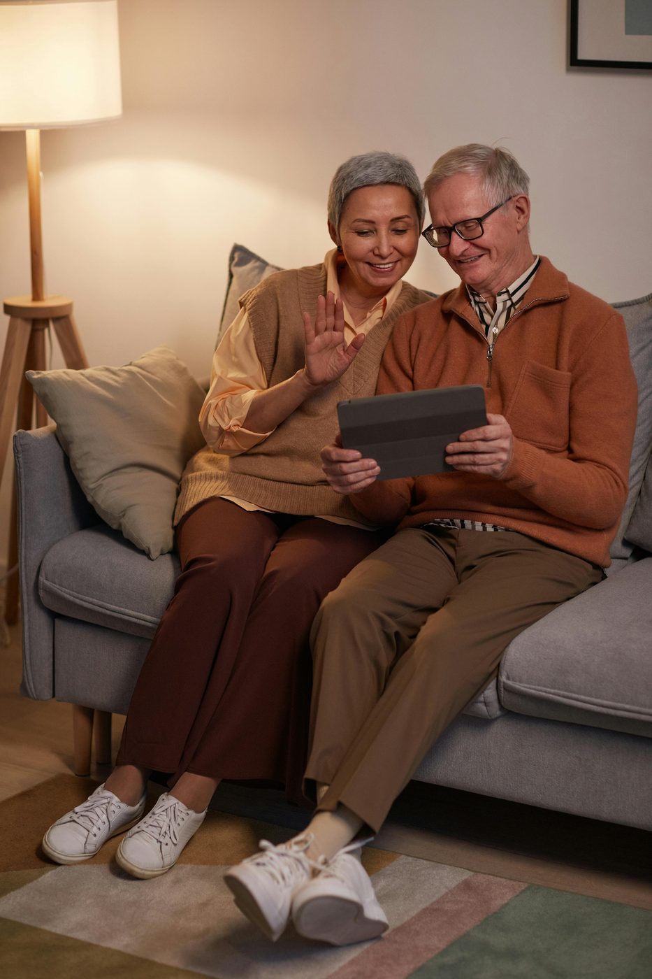 A senior couple smiling and waving at a tablet during a video call from their living room